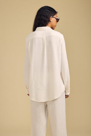 A person wearing the DENISE SHIRT by Velvet by Graham & Spencer—an off-white, textured cotton gauze piece—paired with matching pants, stands with their back to the camera against a beige background.