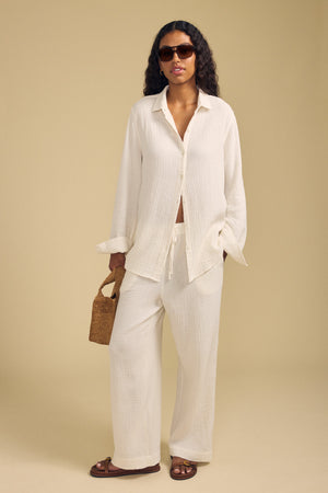 A woman stands against a beige background, wearing Velvet by Graham & Spencer's FRANNY PANT—wide-leg cotton gauze pants with a comfortable pull-on waistband—paired with a loose cream shirt, brown sandals, sunglasses, and a small woven handbag.