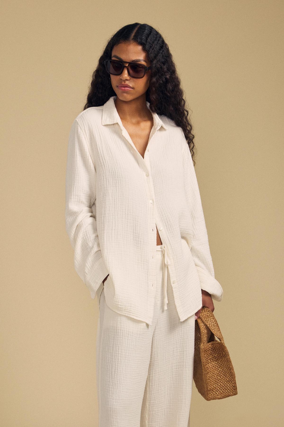 A woman wearing the DENISE SHIRT by Velvet by Graham & Spencer, paired with matching pants and sunglasses, holds a woven brown handbag against a beige background.