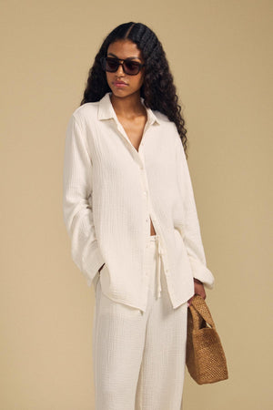 A woman wearing the DENISE SHIRT by Velvet by Graham & Spencer, paired with matching pants and sunglasses, holds a woven brown handbag against a beige background.