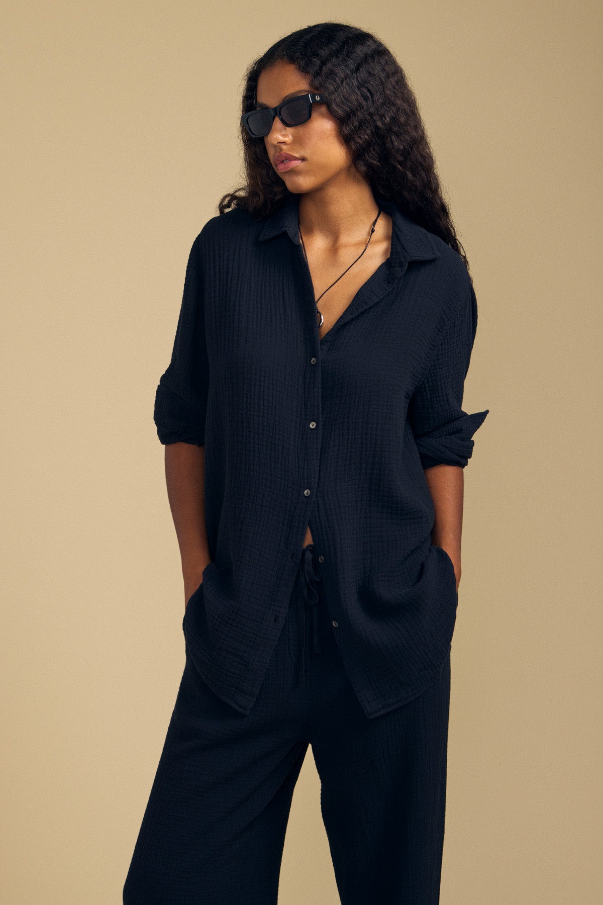 A woman with long curly hair and black sunglasses poses against a neutral background, wearing the DENISE SHIRT from Velvet by Graham & Spencer—a loose, black cotton gauze button-up—paired with matching pants, her hands in her pockets.