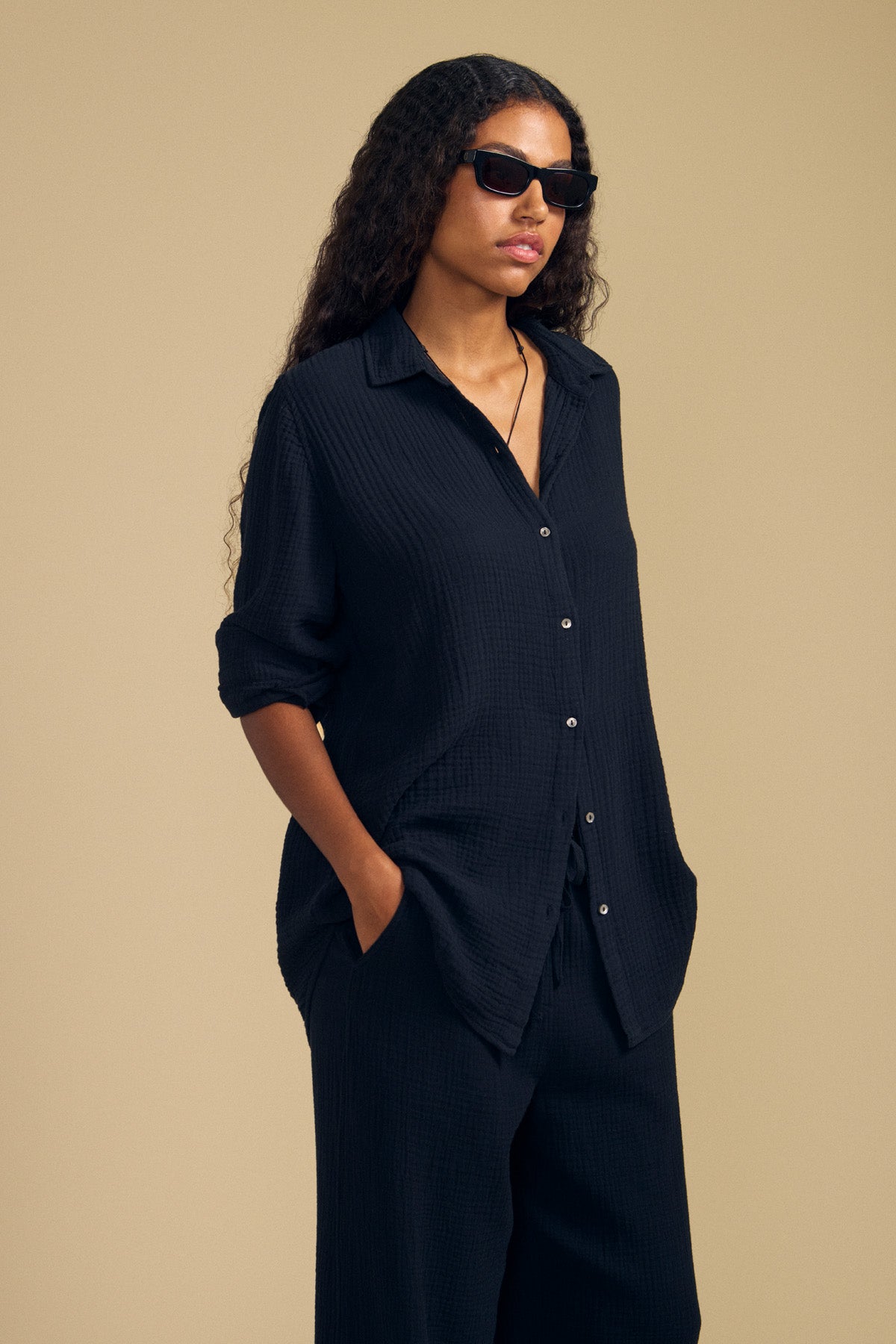 A person with long curly hair and sunglasses stands against a plain beige background, hands in pockets, wearing the DENISE SHIRT by Velvet by Graham & Spencer—a black cotton gauze button-up paired with matching pants. The lightweight gauze fabric lends subtle texture to the look.-41779839041729