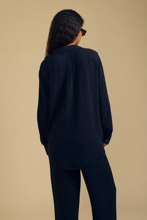 A person with long curly hair wears the Velvet by Graham & Spencer DENISE SHIRT—a loose-fitting black cotton gauze top—paired with matching pants, standing with their back to the camera against a beige background.