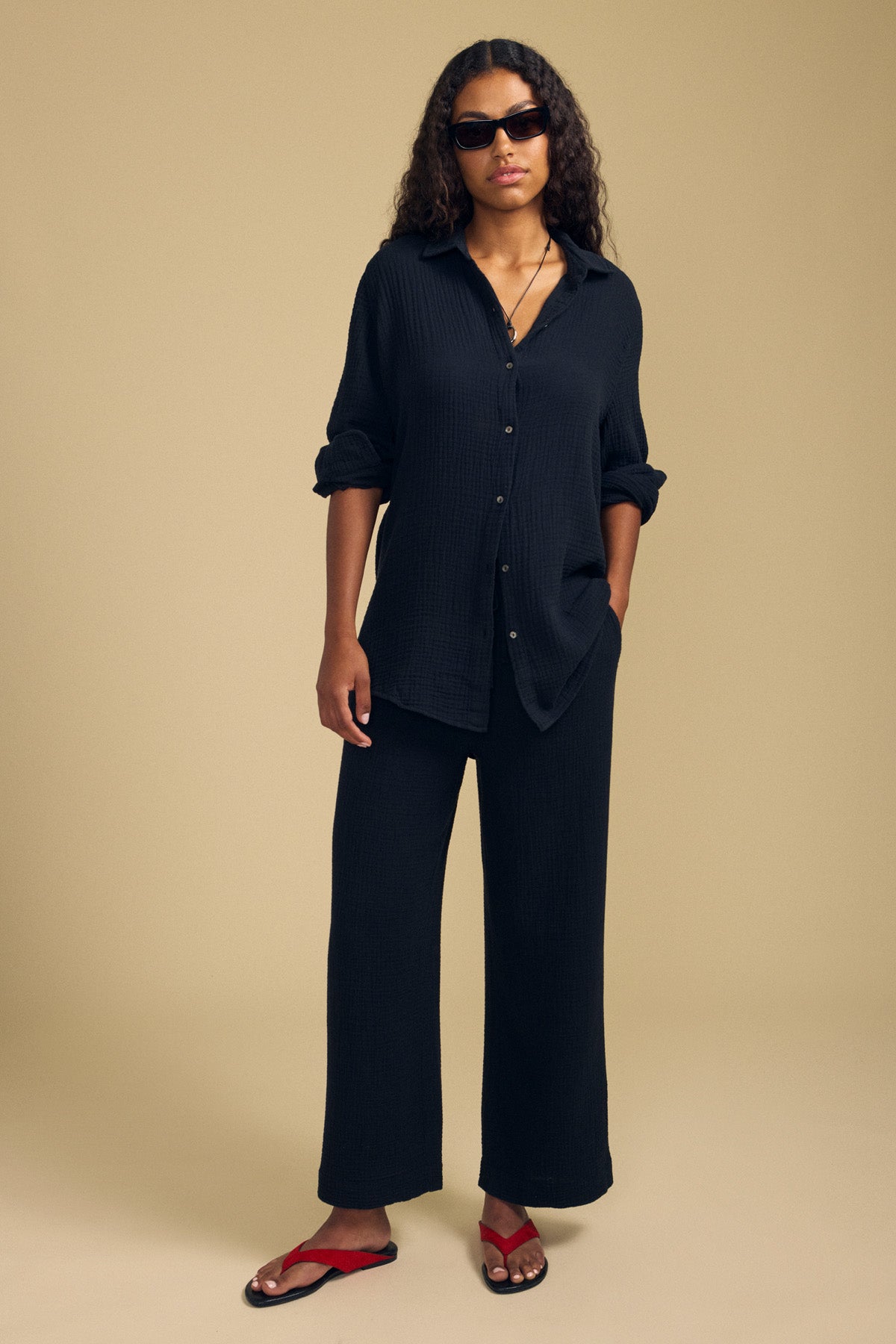 A person wearing a loose black button-up shirt, Velvet by Graham & Spencer’s FRANNY PANT in matching cotton gauze with a comfortable pull-on waistband, red sandals, and sunglasses stands against a plain beige background.-41779846676673