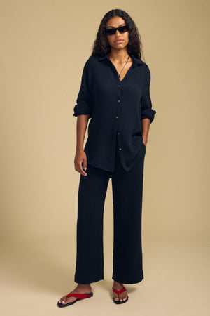 A person wearing a loose black button-up shirt, Velvet by Graham & Spencer’s FRANNY PANT in matching cotton gauze with a comfortable pull-on waistband, red sandals, and sunglasses stands against a plain beige background.