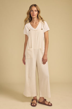 A woman stands against a beige background in sandals, wearing the EVERLEE JUMPSUIT by Velvet by Graham & Spencer—a cream-colored cotton gauze jumpsuit with wide legs and front pockets—layered over a white t-shirt.