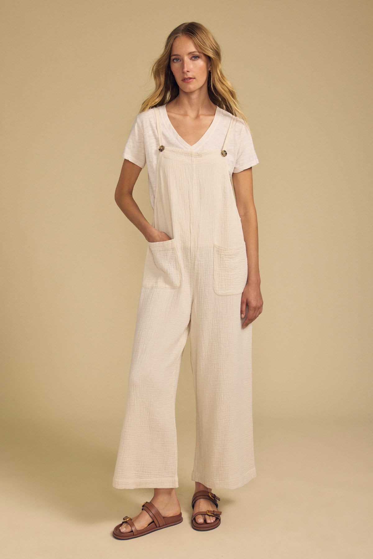 A woman stands against a beige background, wearing the EVERLEE JUMPSUIT by Velvet by Graham & Spencer—a cream cotton gauze wide-leg jumpsuit with front pockets—layered over a white V-neck t-shirt and finished with brown sandals.