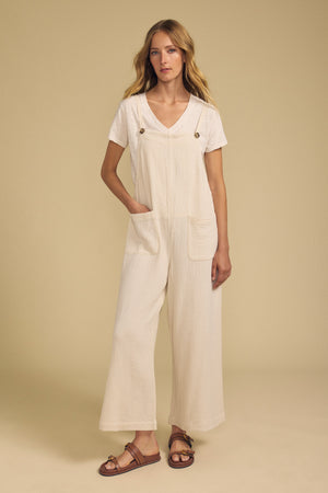 A woman stands against a beige background, wearing the EVERLEE JUMPSUIT by Velvet by Graham & Spencer—a cream cotton gauze wide-leg jumpsuit with front pockets—layered over a white V-neck t-shirt and finished with brown sandals.
