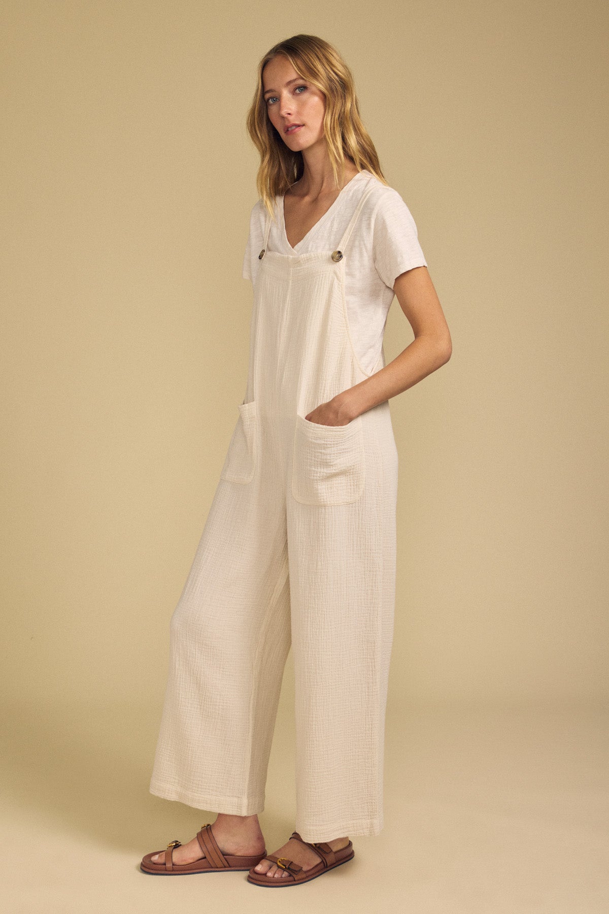 A woman stands against a beige background wearing a white t-shirt, the EVERLEE JUMPSUIT by Velvet by Graham & Spencer—a casual style featuring large pockets and wide legs in a soft cream hue—and brown sandals.-41752187502785