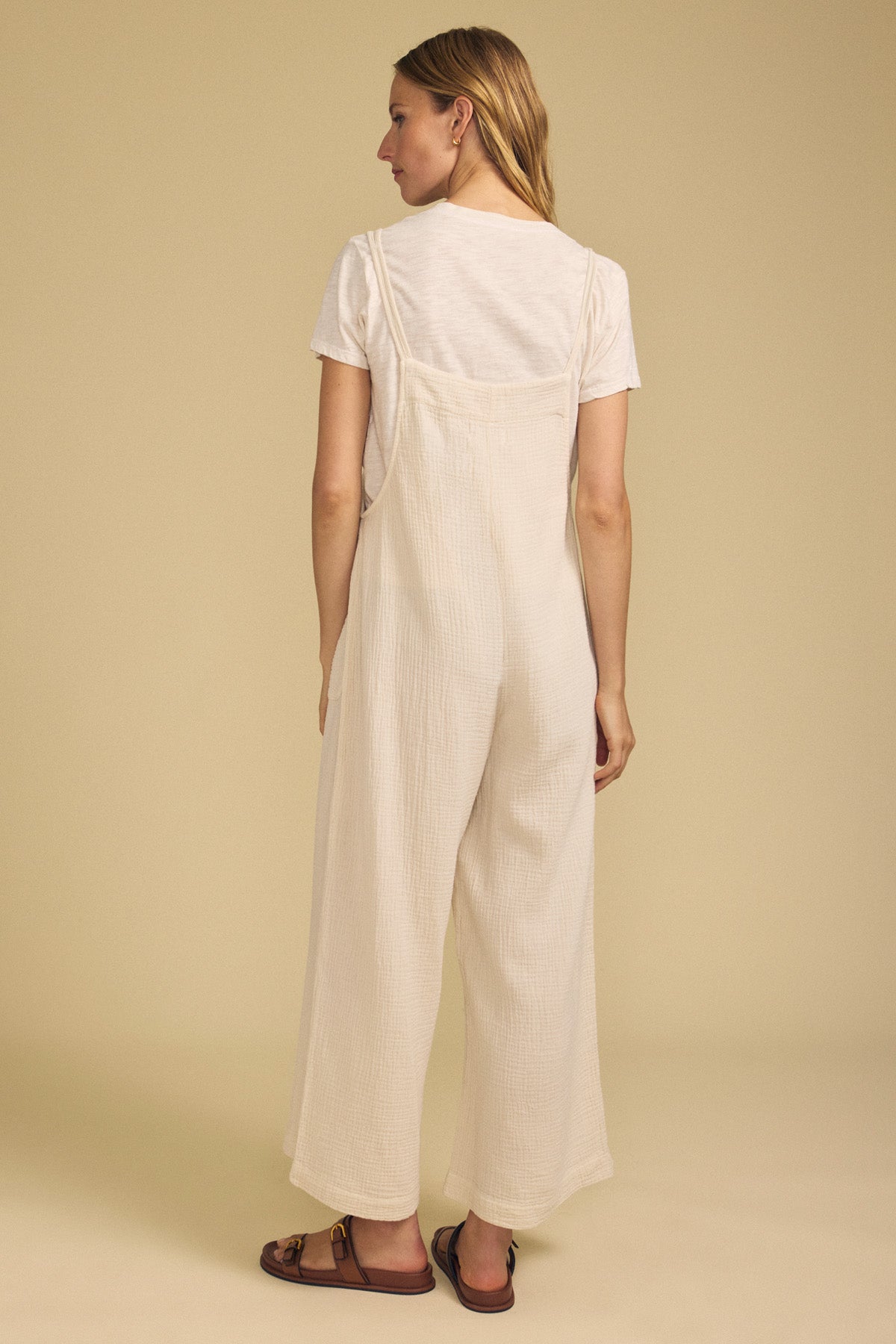 A woman stands facing away, wearing a cream short-sleeve top paired with the EVERLEE JUMPSUIT by Velvet by Graham & Spencer—a loose, wide-leg style—along with brown sandals, against a plain beige background.