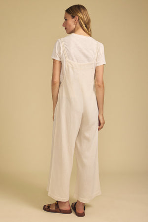 A woman stands facing away, wearing a cream short-sleeve top paired with the EVERLEE JUMPSUIT by Velvet by Graham & Spencer—a loose, wide-leg style—along with brown sandals, against a plain beige background.