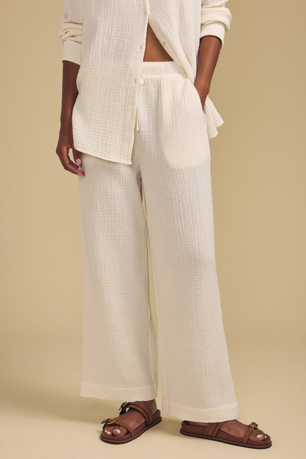 A person wearing the FRANNY PANT by Velvet by Graham & Spencer, a wide leg cotton gauze pant set in a cream tone, paired with brown sandals. Only the lower body and part of the torso are visible as they stand against a beige background.