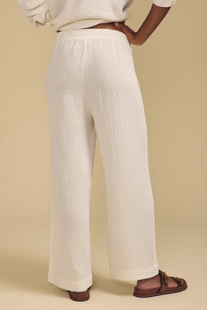 A person wearing the FRANNY PANT by Velvet by Graham & Spencer—loose white cotton gauze pants with a pull-on waistband—paired with a white top and brown sandals, standing against a plain beige background.