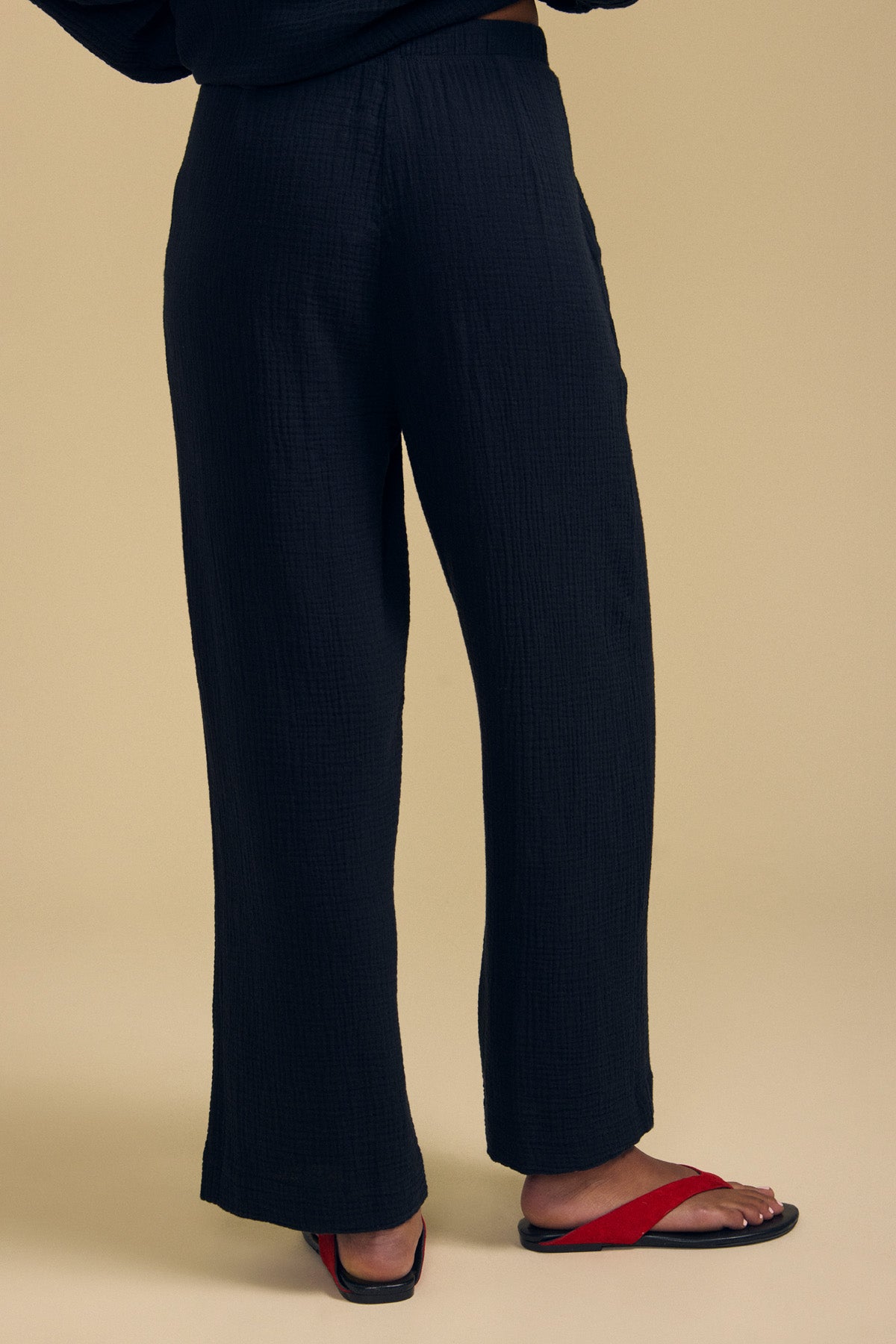 A person is shown from the waist down, wearing the FRANNY PANT by Velvet by Graham & Spencer—loose, wide-leg black pants featuring a comfortable pull-on waistband—paired with red flip-flops against a plain beige background.