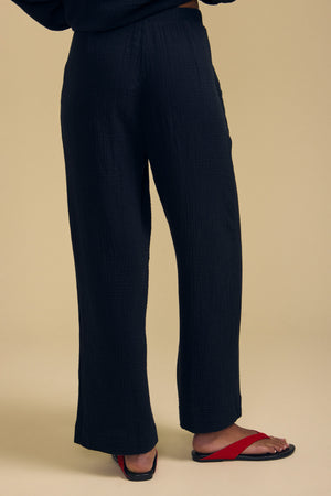 A person is shown from the waist down, wearing the FRANNY PANT by Velvet by Graham & Spencer—loose, wide-leg black pants featuring a comfortable pull-on waistband—paired with red flip-flops against a plain beige background.