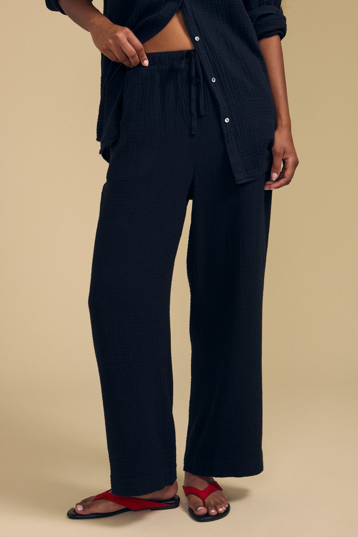A person stands against a beige background wearing the FRANNY PANT by Velvet by Graham & Spencer, featuring loose black cotton gauze with a comfortable pull-on waistband. They pair it with a matching black shirt and red sandals.