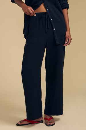 A person stands against a beige background wearing the FRANNY PANT by Velvet by Graham & Spencer, featuring loose black cotton gauze with a comfortable pull-on waistband. They pair it with a matching black shirt and red sandals.