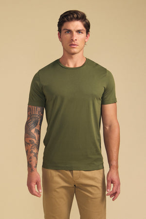 A man with a tattoo on his left arm wears the HOWARD TEE by Velvet by Graham & Spencer in olive green and beige pants, standing against a plain beige background for a vintage feel.
