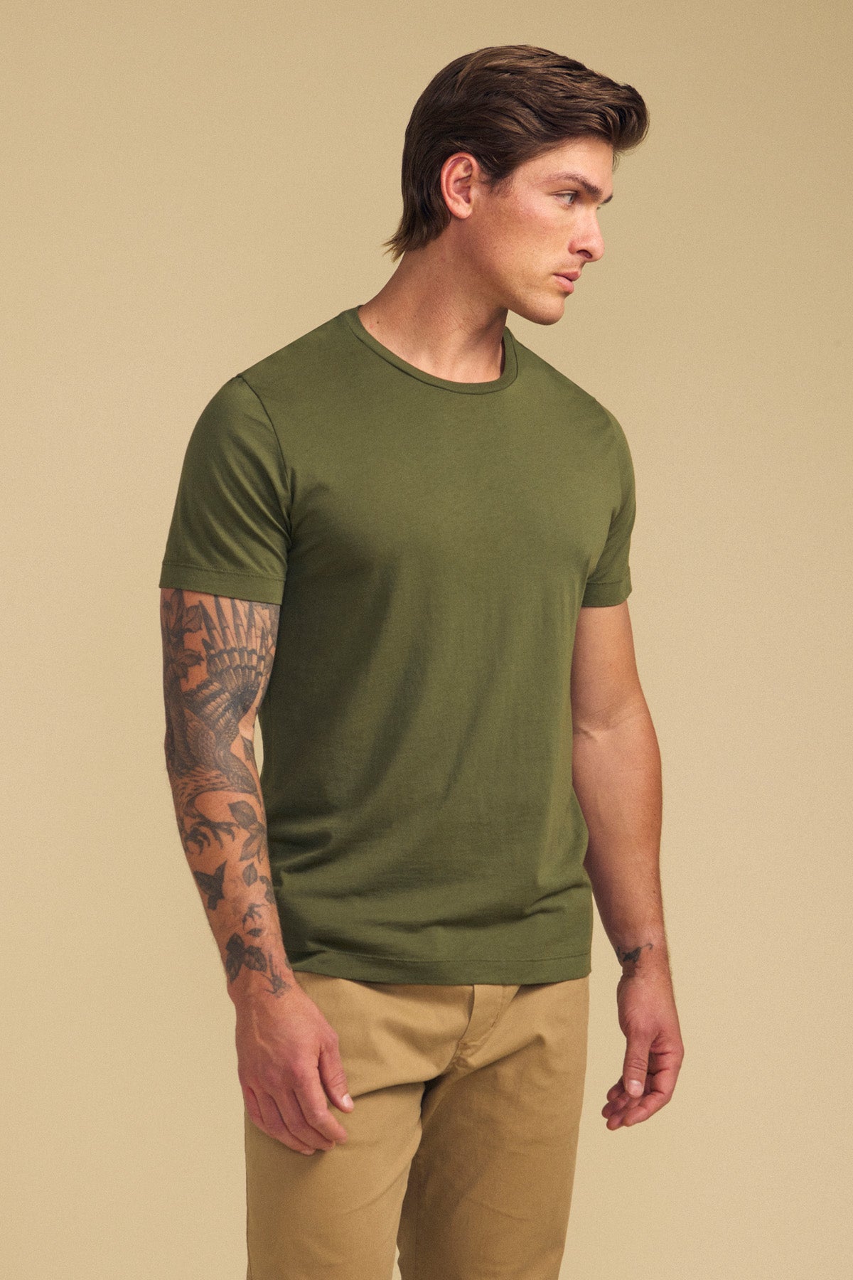 A man with tattoos on his left arm wears the olive green HOWARD TEE by Velvet by Graham & Spencer and tan pants, standing against a beige background and looking to the side.-41702025887937
