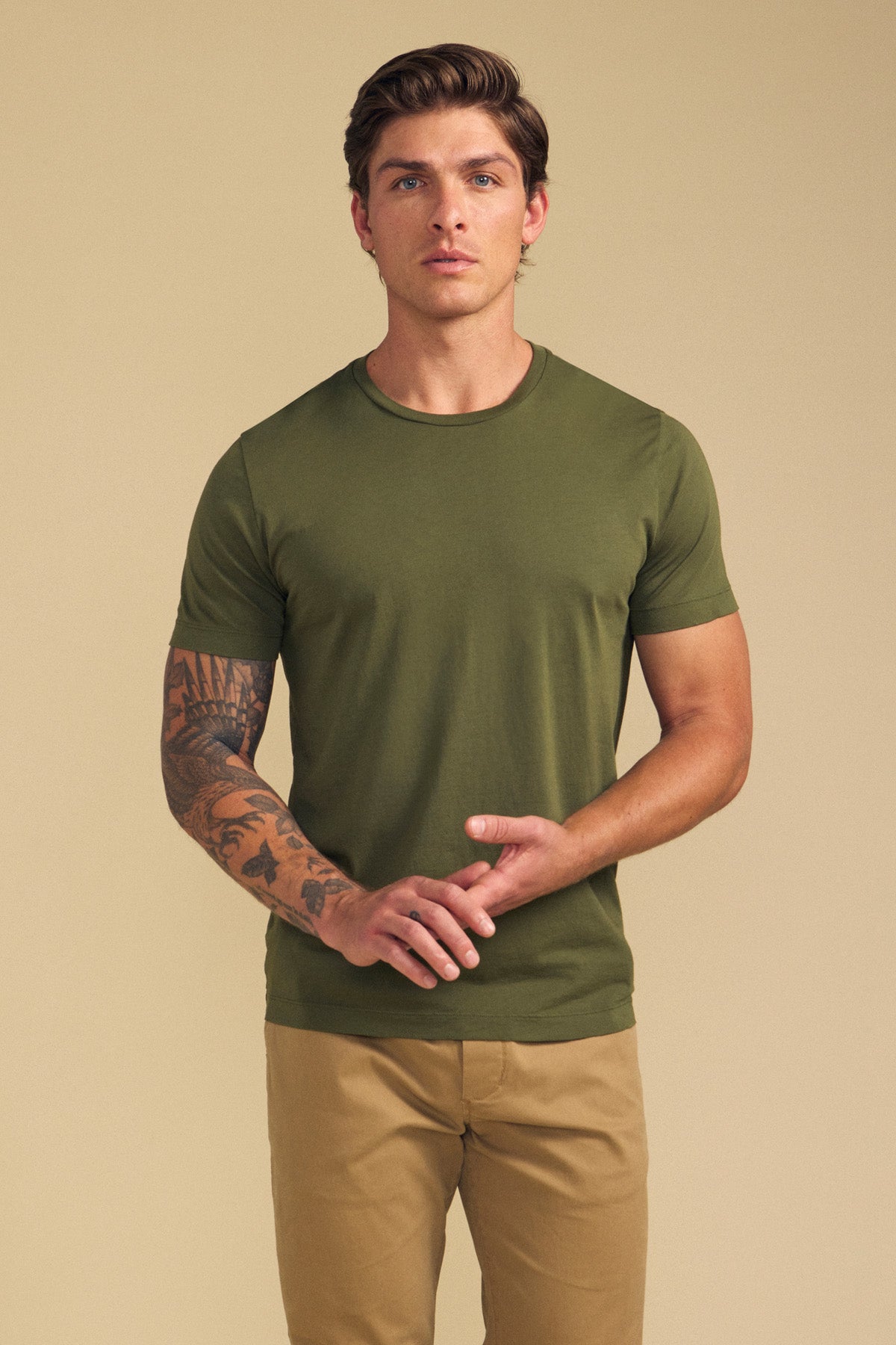 A man models the HOWARD TEE by Velvet by Graham & Spencer in green with tan pants, standing against a beige background. His left forearm tattoo and the tee's perfect fit are clearly visible.