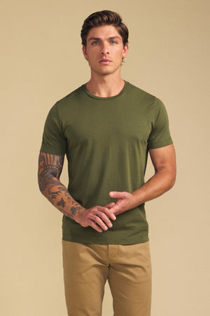 A man models the HOWARD TEE by Velvet by Graham & Spencer in green with tan pants, standing against a beige background. His left forearm tattoo and the tee's perfect fit are clearly visible.