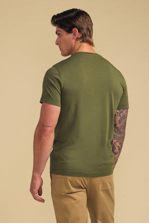 A man faces away, wearing the olive green HOWARD TEE by Velvet by Graham & Spencer and khaki pants. His right arm features a small tattoo; his left, a larger floral tattoo. The neutral background adds a vintage touch.