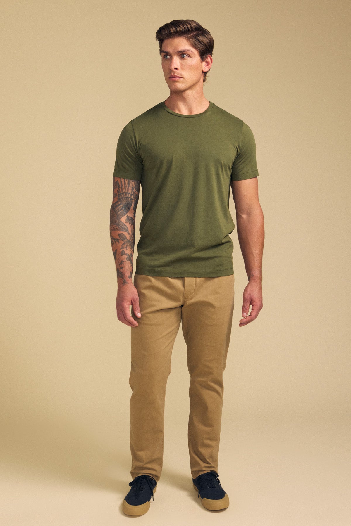 A man faces forward in the HOWARD TEE by Velvet by Graham & Spencer—an olive green Pima jersey short-sleeve shirt—paired with tan pants and black shoes, showing a tattoo sleeve on his left arm against a beige background.