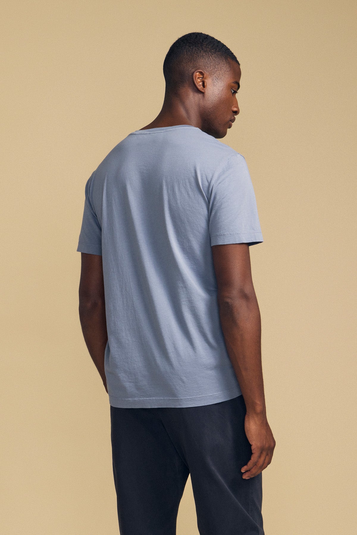 A person stands facing away from the camera, wearing the Velvet by Graham & Spencer HOWARD TEE—a light blue short-sleeve t-shirt made from lightweight pima jersey for a vintage feel and perfect fit—paired with dark pants against a beige background.