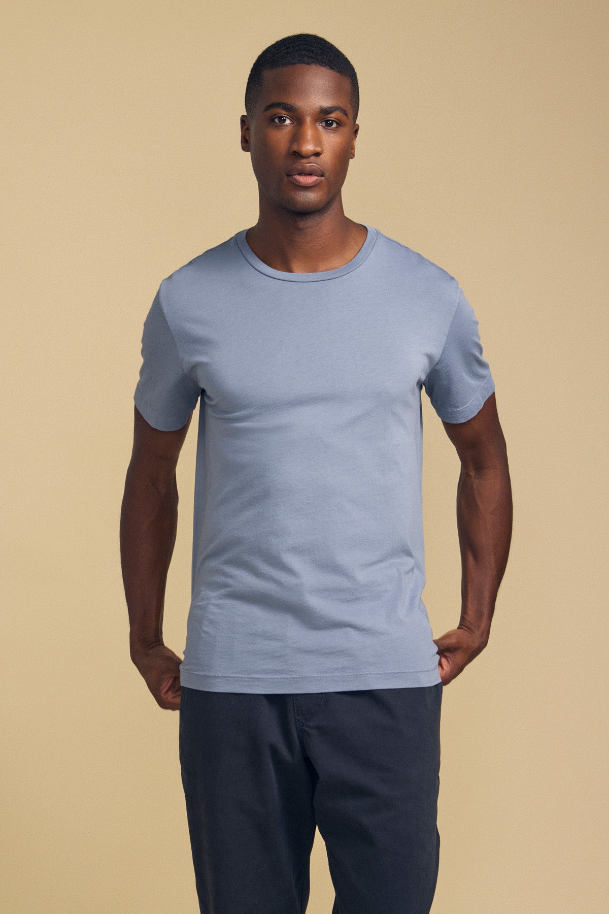 A man wearing the Velvet by Graham & Spencer HOWARD TEE—a light blue, short-sleeve t-shirt in premium Pima jersey—paired with dark pants stands facing the camera against a plain beige background; the tee features a perfect fit and vintage feel.
