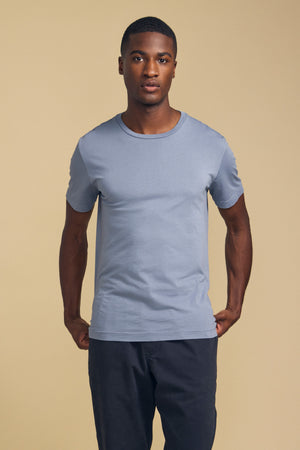 A man wearing the Velvet by Graham & Spencer HOWARD TEE—a light blue, short-sleeve t-shirt in premium Pima jersey—paired with dark pants stands facing the camera against a plain beige background; the tee features a perfect fit and vintage feel.