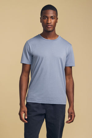 A person stands facing forward, wearing the HOWARD TEE by Velvet by Graham & Spencer—a light blue short-sleeve t-shirt made from lightweight pima jersey for the perfect fit—paired with dark pants against a plain beige background.