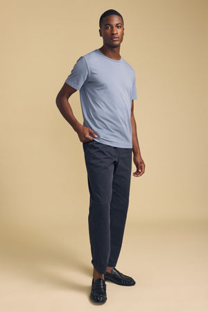 A person stands against a beige background wearing the HOWARD TEE by Velvet by Graham & Spencer—a light blue t-shirt crafted from lightweight pima jersey—paired with dark pants and black loafers, with one hand in their pocket.