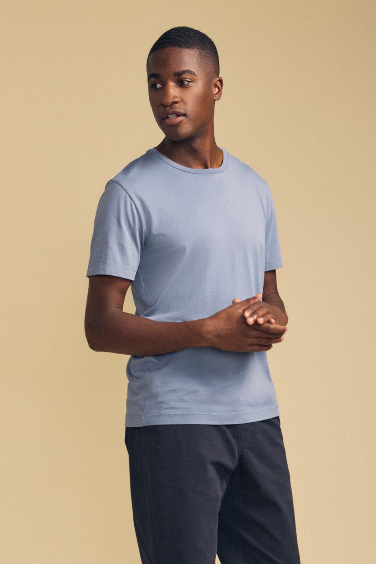A man wearing the HOWARD TEE by Velvet by Graham & Spencer—crafted from lightweight pima jersey in a light blue shade—pairs it with dark pants and stands against a beige background, looking slightly to the side with his hands clasped. The tee’s perfect fit and vintage feel add effortless style to his look.