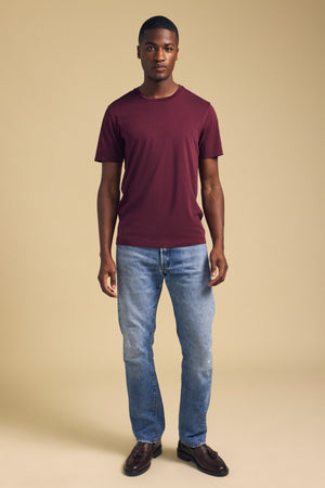 A man wearing a burgundy Velvet by Graham & Spencer HOWARD TEE, light-wash blue jeans, and dark brown leather loafers with a perfect fit.