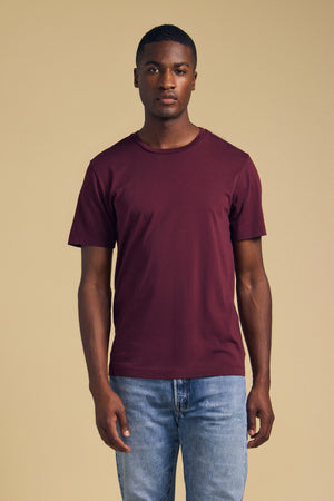 Front view of a dark plum short-sleeve T-shirt showcasing its crew neck on a model against a tan background