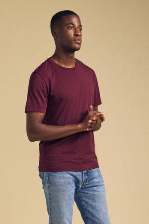 A man wearing a maroon short-sleeved Velvet by Graham & Spencer HOWARD TEE and blue jeans with a vintage feel.