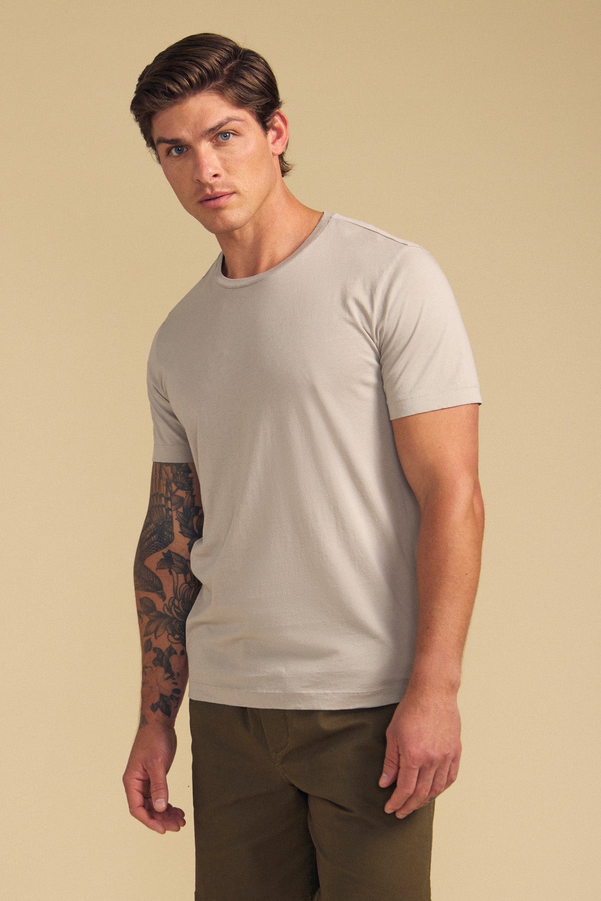 A man with short brown hair and a tattoo sleeve models the Velvet by Graham & Spencer HOWARD TEE, a lightweight beige Pima jersey t-shirt, paired with olive pants against a beige background, highlighting its perfect fit.