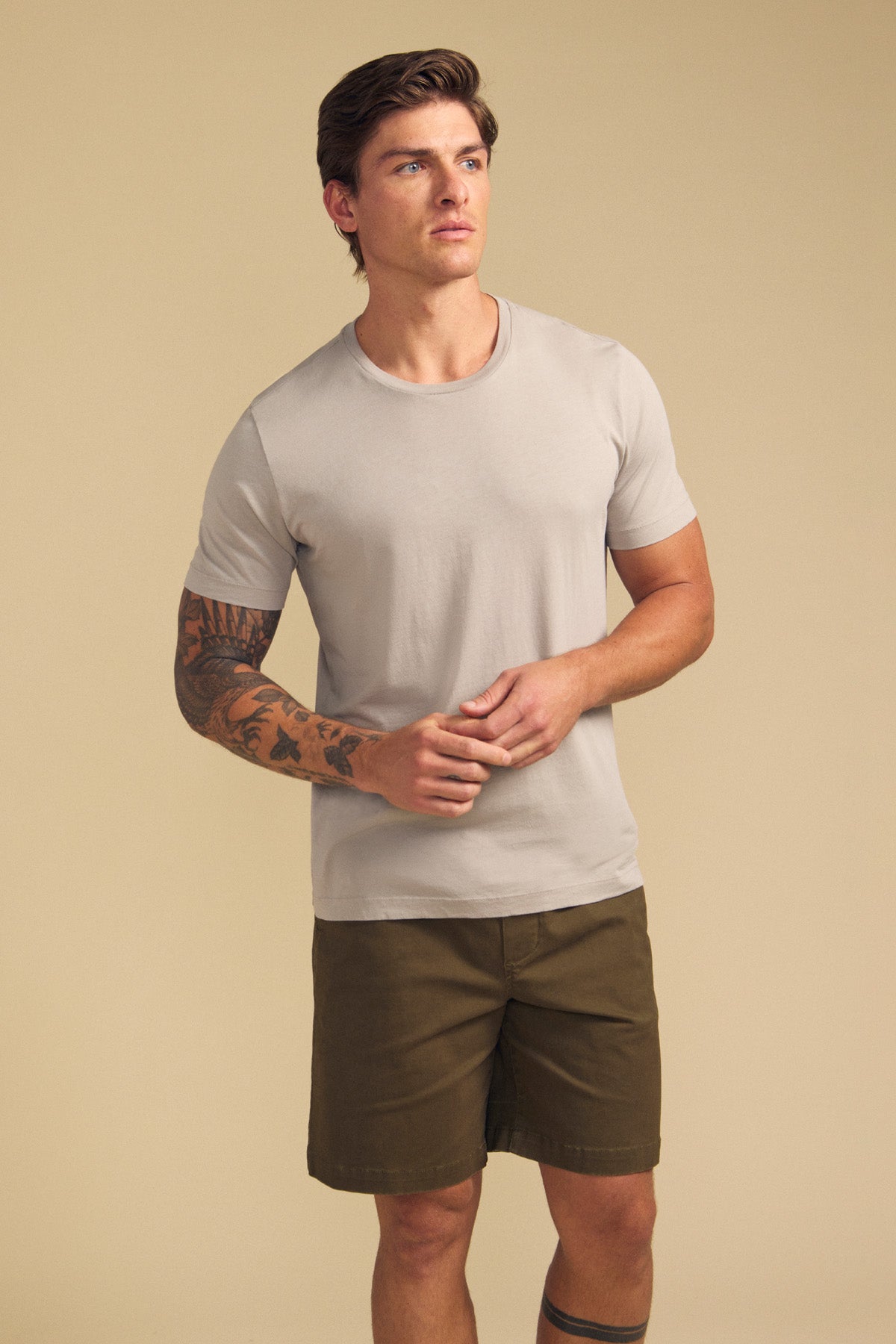 A man stands against a beige background, wearing Velvet by Graham & Spencer’s light gray HOWARD TEE and olive green shorts. He has a tattoo on his left arm and looks off to the side.-41702147588289