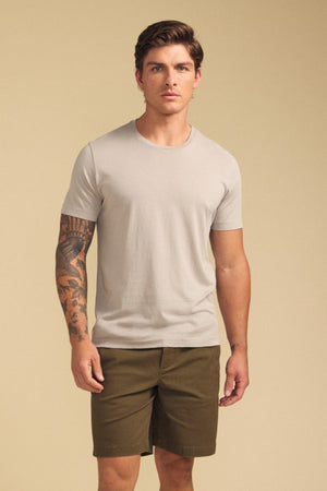A man with a tattooed left arm wears the Velvet by Graham & Spencer HOWARD TEE in light beige and olive green shorts, standing against a plain beige background.