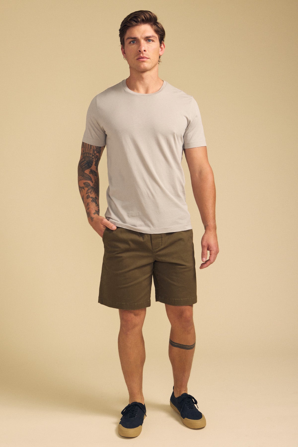 A man with tattoos on his left arm and right leg stands against a beige background wearing the Velvet by Graham & Spencer HOWARD TEE in light gray, olive green shorts, and dark sneakers.