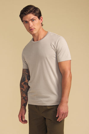 A man with short brown hair and a tattoo sleeve models the Velvet by Graham & Spencer HOWARD TEE, a lightweight beige Pima jersey t-shirt, paired with olive pants against a beige background, highlighting its perfect fit.