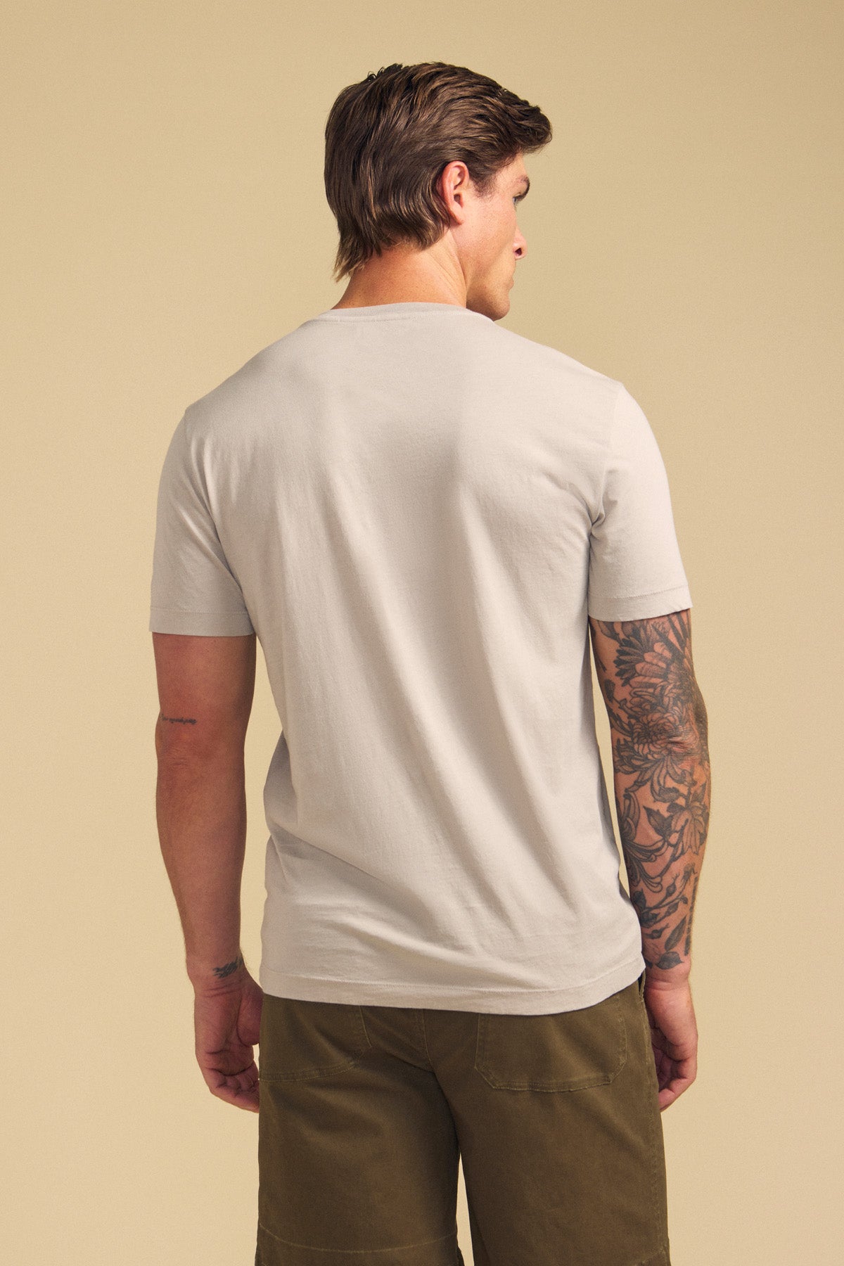 A man faces away, wearing the Velvet by Graham & Spencer HOWARD TEE in light gray Pima jersey and olive green shorts. His right arm has a tattoo sleeve. Background is plain beige.-41702147719361