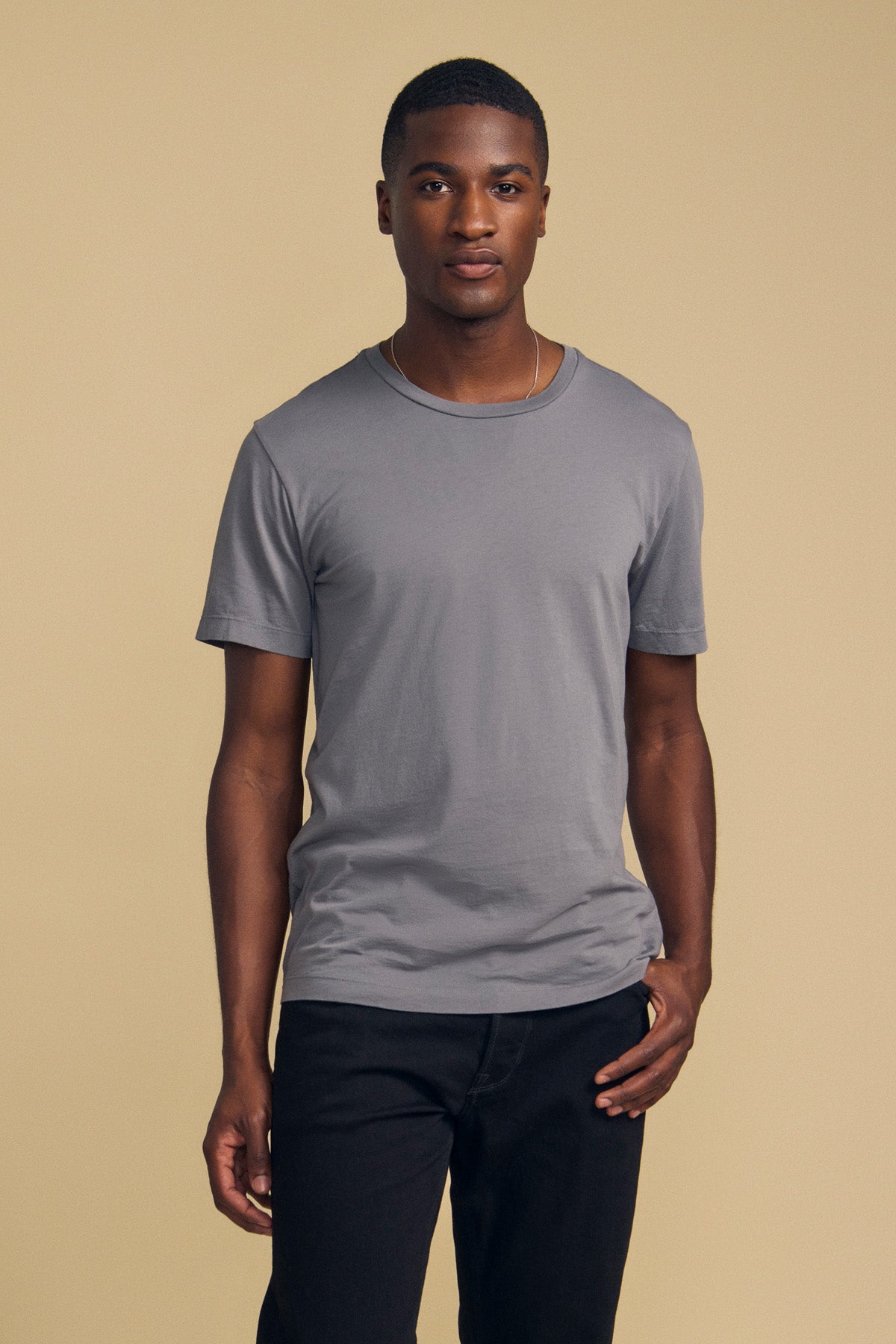 A person stands against a beige background, wearing the HOWARD TEE by Velvet by Graham & Spencer—an ultra-soft Pima jersey short-sleeve t-shirt in gray designed for a vintage feel and perfect fit—paired with black pants.-41859668246721