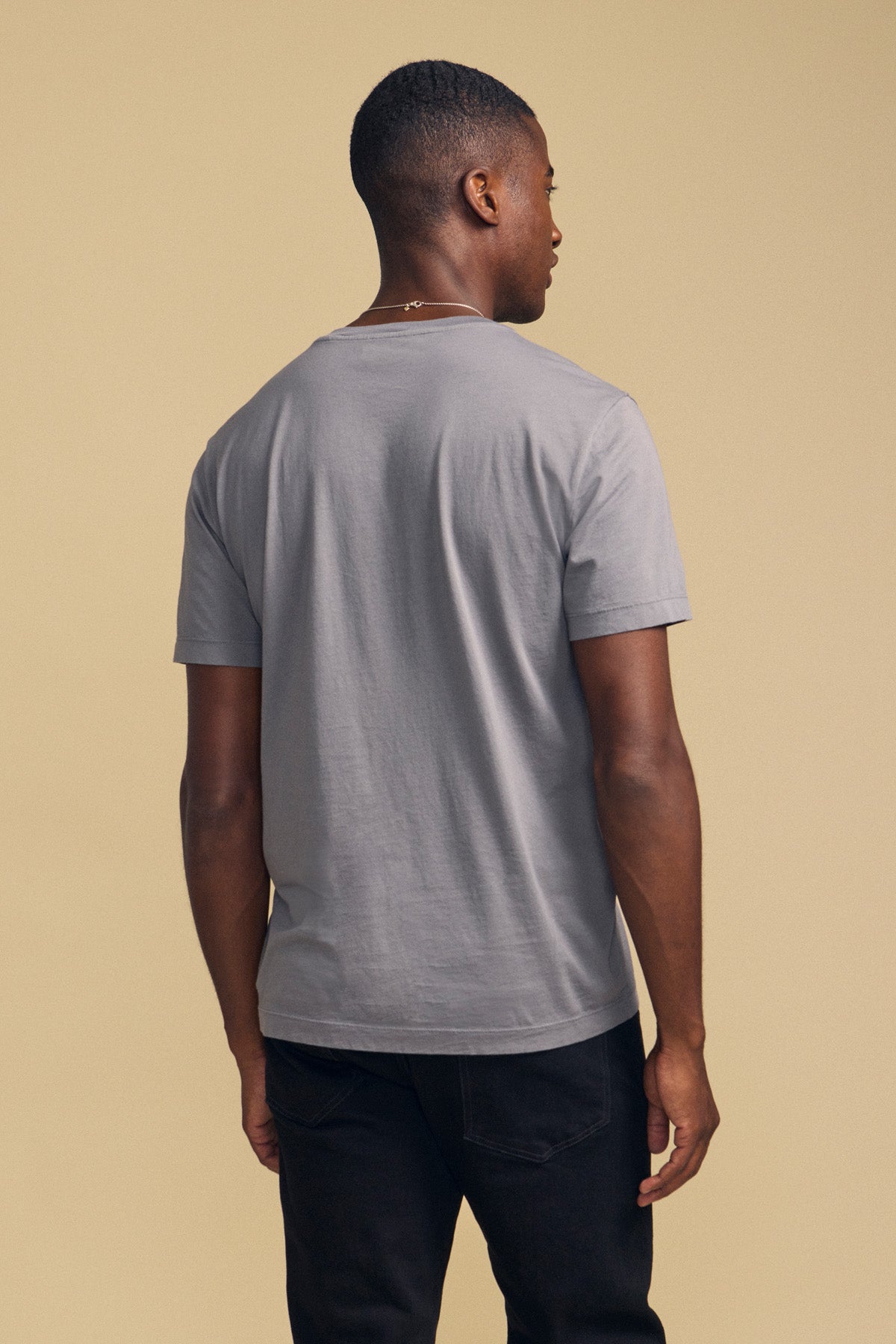 A man stands facing away from the camera against a beige background, wearing the HOWARD TEE by Velvet by Graham & Spencer—a plain light gray Pima jersey t-shirt paired with black jeans—showcasing a perfect fit and subtle vintage feel.