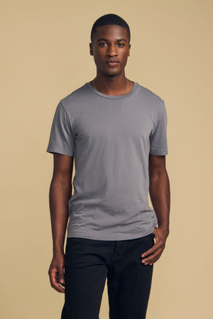 A person stands against a beige background, wearing the HOWARD TEE by Velvet by Graham & Spencer—an ultra-soft Pima jersey short-sleeve t-shirt in gray designed for a vintage feel and perfect fit—paired with black pants.