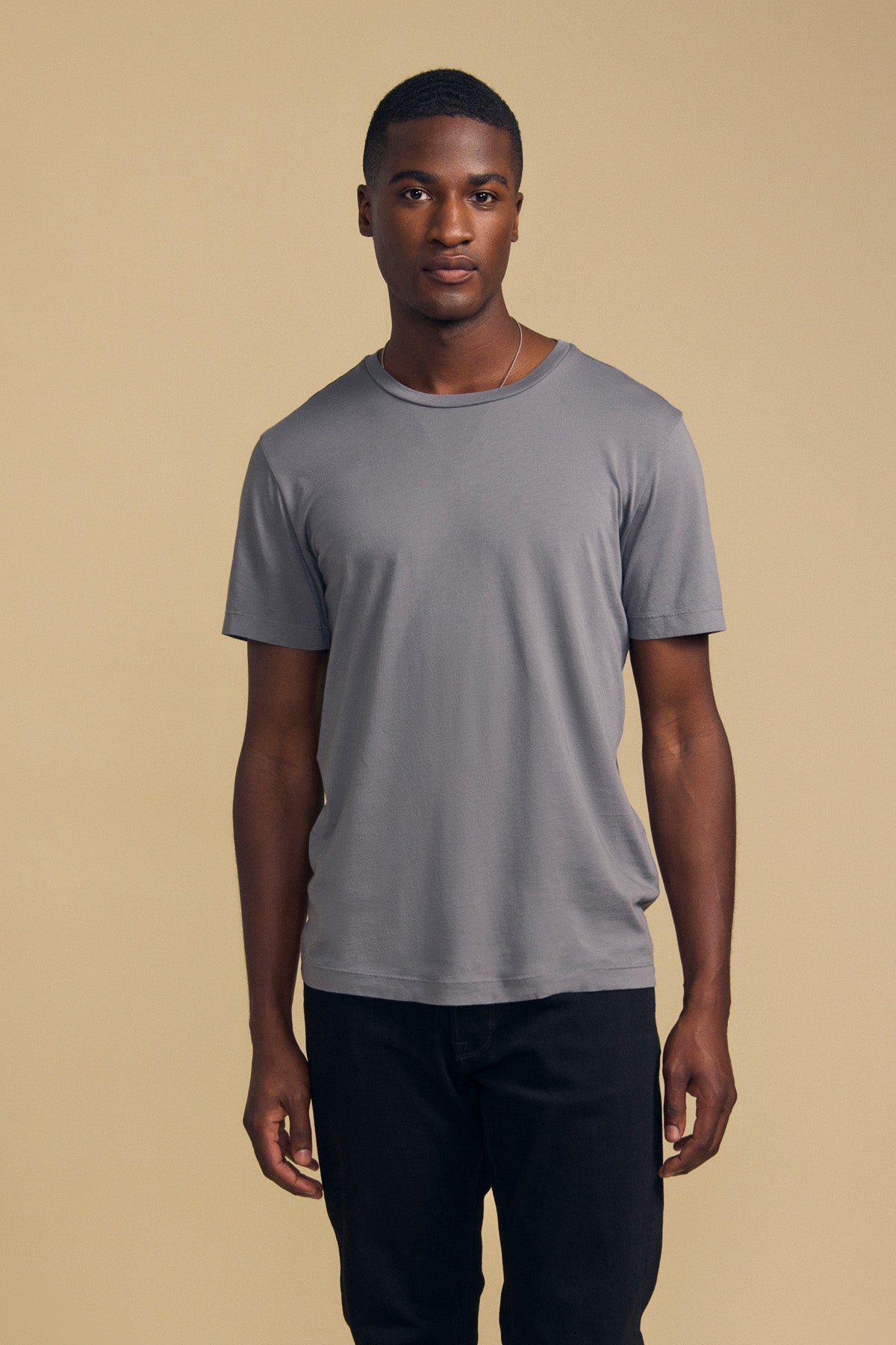 A man stands facing forward against a beige background, wearing the Velvet by Graham & Spencer HOWARD TEE—a plain gray short-sleeve Pima jersey t-shirt—and black pants.-41859668279489
