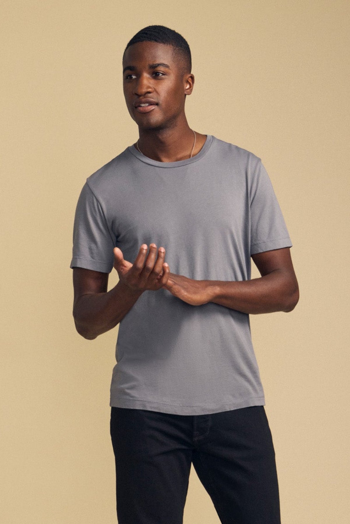 A man stands against a beige background, wearing the HOWARD TEE—a gray short-sleeve Pima jersey t-shirt by Velvet by Graham & Spencer—paired with perfectly fitting black pants, as he gestures with his hands in front of him.-41859668377793