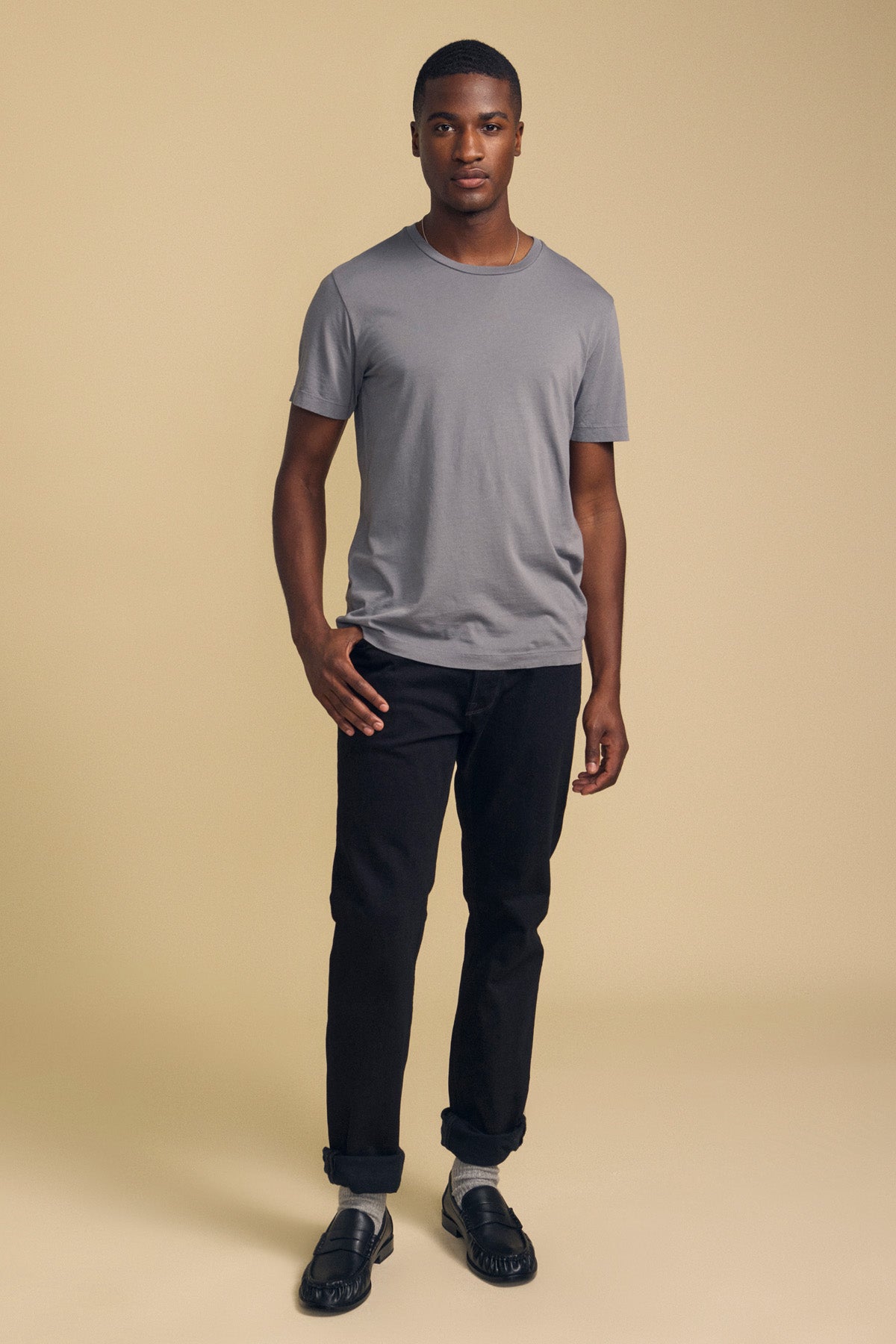 A man stands facing forward against a beige background, wearing the HOWARD TEE by Velvet by Graham & Spencer—a gray Pima jersey t-shirt with a vintage feel—paired with cuffed black jeans for the perfect fit and black shoes.-41859668443329