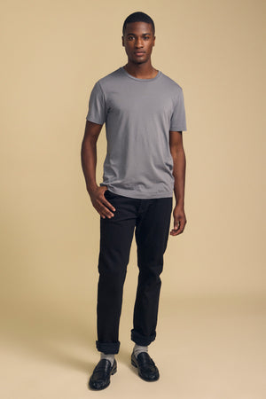 A man stands facing forward against a beige background, wearing the HOWARD TEE by Velvet by Graham & Spencer—a gray Pima jersey t-shirt with a vintage feel—paired with cuffed black jeans for the perfect fit and black shoes.