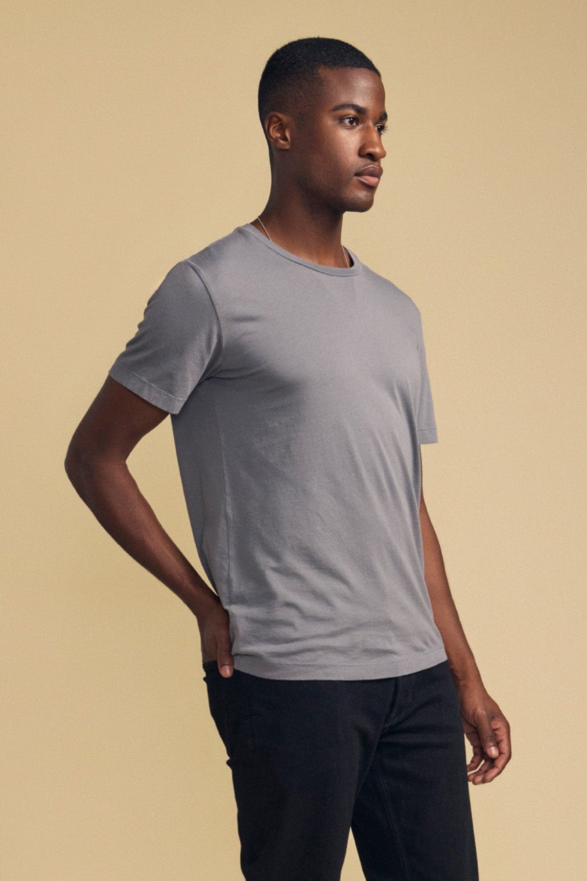 A man stands against a beige background, wearing Velvet by Graham & Spencer’s HOWARD TEE, a plain grey short-sleeve Pima jersey t-shirt, paired with black pants and resting one hand on his hip.-41859668508865